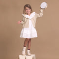 A young girl in a white dress and beige cardigan stands on a wooden block, smiling and holding a tambourine above her head, wearing Pretty Brave Girls Maddison Winter Bloom Pink Leather Mary Jane T Bar shoes against a plain tan background., 9420075362840, 9420075362833, 9420075362826, 9420075362819, 9420075362802, 9420075362796, 50965, 50966, 50967, 50968