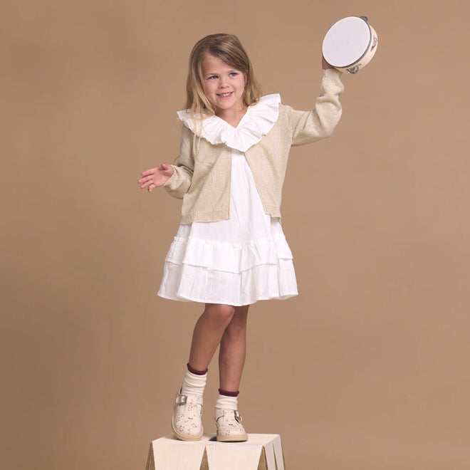 A young girl in a white dress and beige cardigan stands on a wooden block, smiling and holding a tambourine above her head, wearing Pretty Brave Girls Maddison Winter Bloom Pink Leather Mary Jane T Bar shoes against a plain tan background., 9420075362840, 9420075362833, 9420075362826, 9420075362819, 9420075362802, 9420075362796, 50965, 50966, 50967, 50968