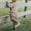 A young child in a beige sweater, khaki pants, and Pretty Braver Blake Leather Boot First Walker Tan stands on grass holding a wooden fence, looking over it outdoors on a slightly cloudy day., 9420075329140, 9420075329133, 9420075329126