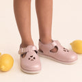 A child wears Pretty Brave Clara Closed Toe Sandal Blush Pearl Pink, standing near yellow lemons on a cream-colored background.