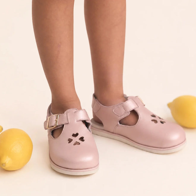 A child wears Pretty Brave Clara Closed Toe Sandal Blush Pearl Pink, standing near yellow lemons on a cream-colored background.