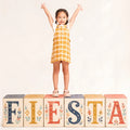 Young girl in a yellow plaid dress stands on FIESTA blocks, wearing Pretty Brave Clara Closed Toe Sandal Blush Pearl Pink, arms raised and smiling against a plain light background.