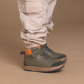 A child wears light beige cargo pants and Pretty Brave Hi Top Boot Corey Olive kids leather boots with black soles and an orange heel accent, standing against a tan background., 50474, 50475, 9420075364370, 9420075364363, 9420075364356, 9420075364349, 9420075364332, 9420075364325, 9420075364318, 9420075364301, 50484, 50485