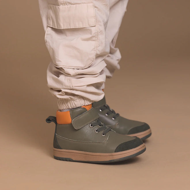 A child wears light beige cargo pants and Pretty Brave Hi Top Boot Corey Olive kids leather boots with black soles and an orange heel accent, standing against a tan background., 50474, 50475, 9420075364370, 9420075364363, 9420075364356, 9420075364349, 9420075364332, 9420075364325, 9420075364318, 9420075364301, 50484, 50485