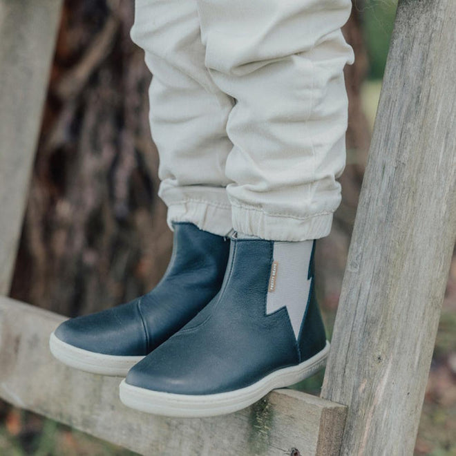 A child in light-colored pants and Pretty Brave Electric Leather Boot - Jet Black, featuring a lightning bolt design and APMA approval, stands outdoors on a wooden fence or ladder., 35922, 9420075327986, 35924, 35925, 9420075327955, 9420075327948, 9420075327931, 9420075327924, 9420075327917, 9420075327894