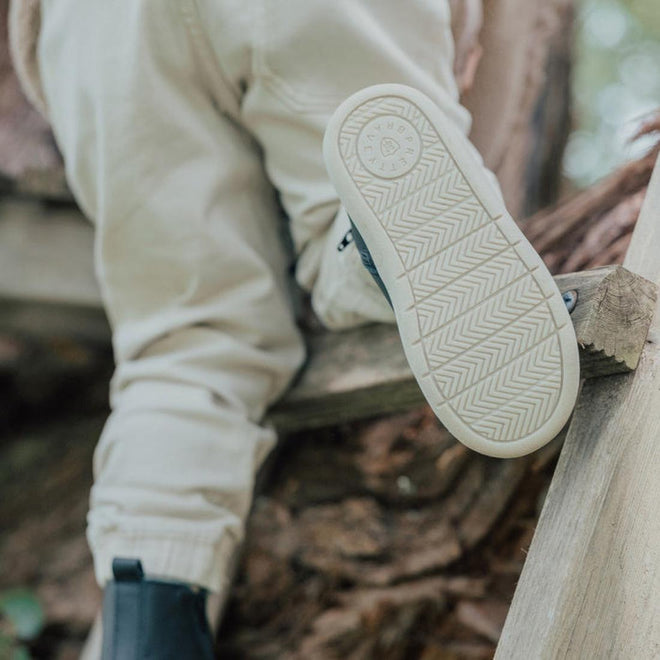 A person in light-colored pants and Pretty Brave Electric Leather Boot - Jet Black sits outdoors on a wooden structure, showing the shoe’s lightning bolt sole design to the camera., 35922, 9420075327986, 35924, 35925, 9420075327955, 9420075327948, 9420075327931, 9420075327924, 9420075327917, 9420075327894