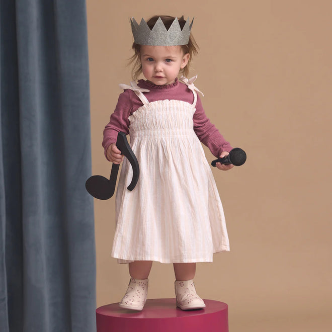 A young child in a paper crown and pink top stands on a round platform holding large musical note props, wearing Pretty Brave Charlie Leather Boot First Walkers Winter Bloom Pink, with a neutral background and blue curtain to the side., 9420075361881, 9420075361874, 9420075361867, 9420075361850