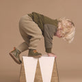 A young child with blonde curly hair wears Pretty Brave Jessie First Walker Hi Top Leather Olive Green shoes while climbing a white and gold triangular block—podiatrist-approved style showcased against a beige background., 9420075361782, 9420075361775, 9420075361768, 9420075361751