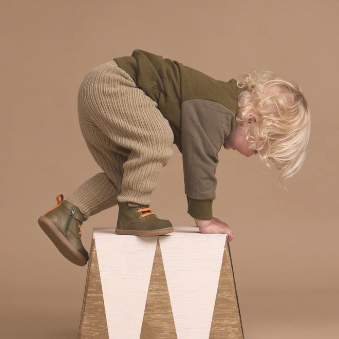 A young child with blonde curly hair wears Pretty Brave Jessie First Walker Hi Top Leather Olive Green shoes while climbing a white and gold triangular block—podiatrist-approved style showcased against a beige background., 9420075361782, 9420075361775, 9420075361768, 9420075361751