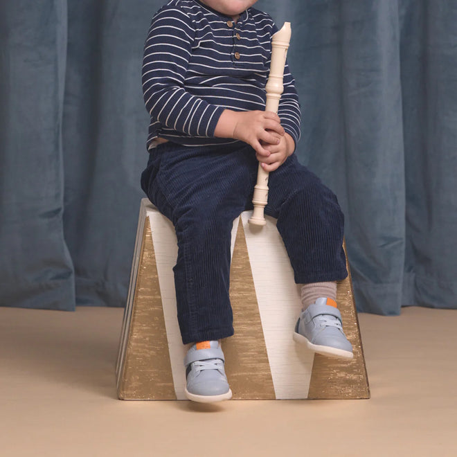 A young child, wearing a navy striped shirt, blue pants, and Pretty Brave SID Leather Trainer First Walker Indigo Blue Grey shoes, sits on a gold and white block holding a white recorder; the child's face is not visible., 9420075362918, 9420075362901, 9420075362895, 9420075362888