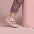 A toddler stands with one foot slightly lifted, wearing mauve pants and Pretty Brave First Walker Toto Leather Trainer shoes in Dusky Pink, beside a light pink cylinder against a soft pink background., 9420075366329, 9420075366312, 9420075366305, 50976