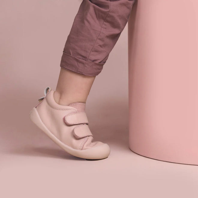 A toddler stands with one foot slightly lifted, wearing mauve pants and Pretty Brave First Walker Toto Leather Trainer shoes in Dusky Pink, beside a light pink cylinder against a soft pink background., 9420075366329, 9420075366312, 9420075366305, 50976