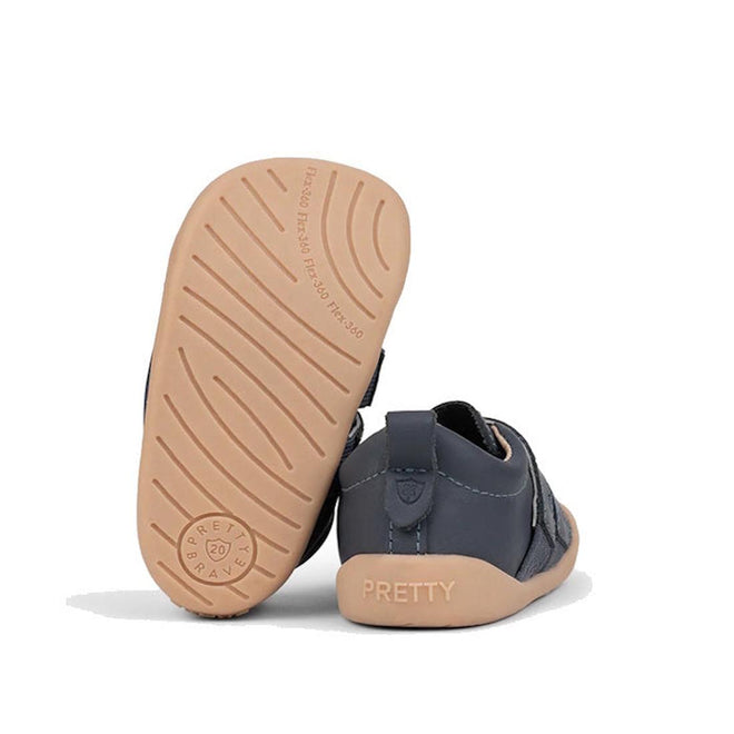 A pair of Pretty Brave First Walker Toto Leather Trainer sneakers in indigo blue leather with light tan flexible rubber soles, featuring curved grooves and "PRETTY BRAVE" stamped on the sole; one shoe stands upright, the other shows the heel., 9420075366275, 9420075366268, 9420075366251, 50971