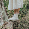 Wearing a white dress and Pretty Brave Electric Leather Boot - Secret Garden, a person stands on a tree stump in lush greenery. Only their lower legs and feet are shown., 45094, 9420075348950, 45096, 9420075348936, 9420075348929, 9420075348912, 9420075348905, 45101, 45102, 45103