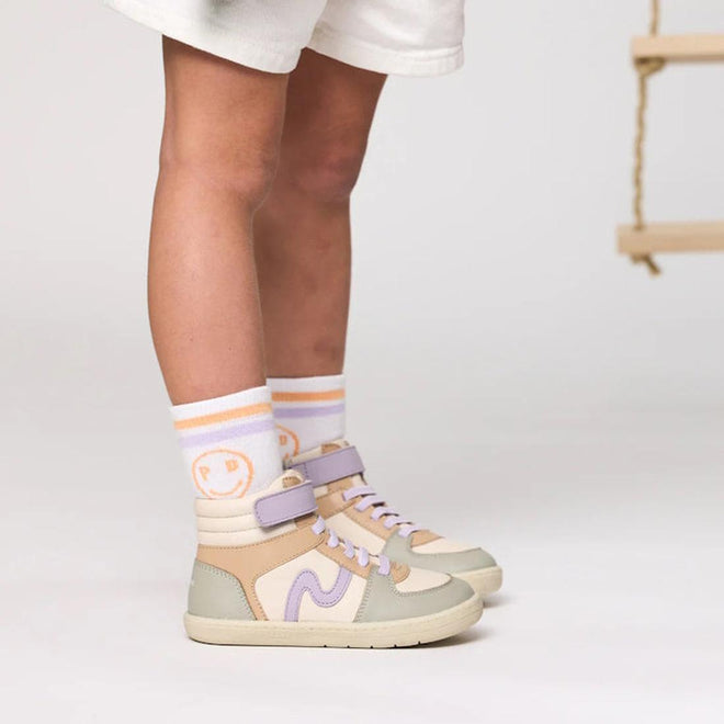 A child wears Pretty Brave Socks 2 Pair Pack Smiley Blush/White with white shorts and cream-lavender high-top sneakers, standing indoors on a light background. A wooden swing appears partially visible to the right., 36744, 36745, 36746