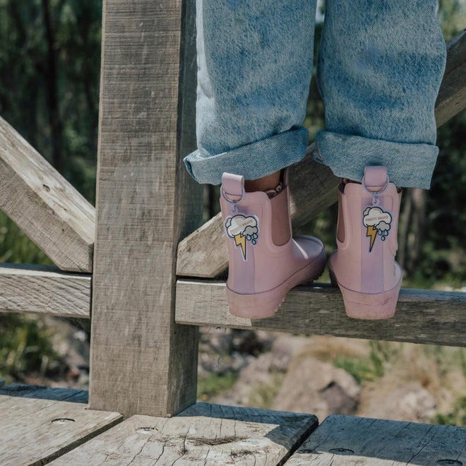 A person wearing rolled-up blue jeans and Pretty Brave Puddle Boot Ankle Rainboot Blush Pink stands on a wooden fence, with green trees and rocks in the background., 9420075328525, 9420075328518, 9420075328501, 9420075328495, 9420075328488, 9420075328471, 9420075328464, 9420075328457, 9420075328440, 9420075328433, 35976
