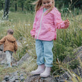 A young girl in a pink jacket and jeans stands on a rock in a grassy area, wearing Pretty Brave Puddle Boot Ankle Rainboot Blush Pink, while another child in a brown jacket walks away. Both kids seem to be exploring nature., 9420075328525, 9420075328518, 9420075328501, 9420075328495, 9420075328488, 9420075328471, 9420075328464, 9420075328457, 9420075328440, 9420075328433, 35976