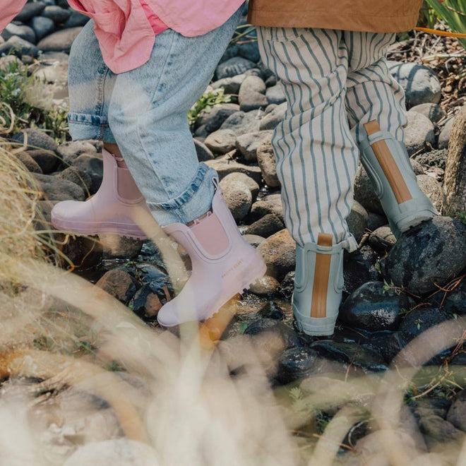 Two children wear casual clothes and stand on wet rocks outdoors. One wears Pretty Brave Puddle Boot Ankle Rainboot Blush Pink and jeans, while the other has gray boots with striped pants. Green plants grow among the rocks., 9420075328525, 9420075328518, 9420075328501, 9420075328495, 9420075328488, 9420075328471, 9420075328464, 9420075328457, 9420075328440, 9420075328433, 35976