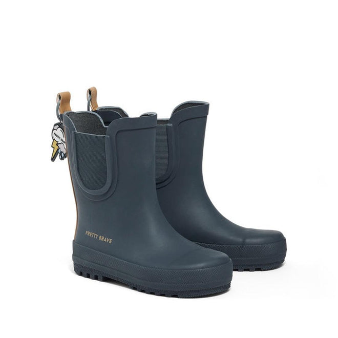 A pair of Pretty Brave Puddle Boot Ankle Rainboot in Muted Indigo Blue with tan pull tabs, a decorative keychain, and "PRETTY BRAVE" in gold on the side. These kids boots are lightweight and fully waterproof.