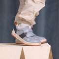A child in light-colored pants and white socks stands on a white wooden surface, wearing Pretty Brave River Bolts Leather Trainer Blue Grey sneakers with podiatrist approval, Velcro straps, and orange accents., 9420075363984, 9420075363977, 9420075363960, 9420075363953, 50510, 50511, 50512, 50513, 50514, 50515, 9420075363984, 9420075363977, 9420075363960, 9420075363953, 9420075363946, 50511, 50512, 50513, 50514, 50515