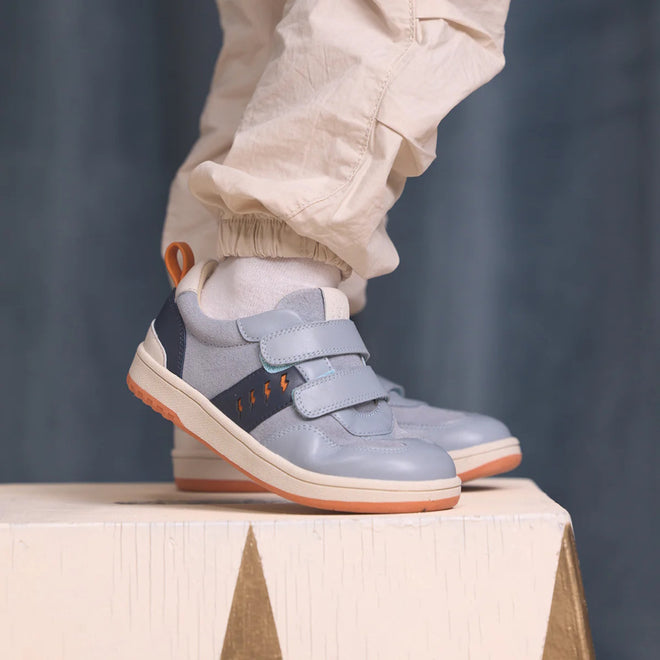A child in light-colored pants and white socks stands on a white wooden surface, wearing Pretty Brave River Bolts Leather Trainer Blue Grey sneakers with podiatrist approval, Velcro straps, and orange accents., 9420075363984, 9420075363977, 9420075363960, 9420075363953, 50510, 50511, 50512, 50513, 50514, 50515, 9420075363984, 9420075363977, 9420075363960, 9420075363953, 9420075363946, 50511, 50512, 50513, 50514, 50515
