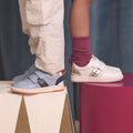 Two children stand on colorful blocks, showing only their legs and feet. One wears Pretty Brave River Bolts Leather Trainer Blue Grey with cream pants; the other wears white sporty shoes with pink socks and a skirt., 9420075363984, 9420075363977, 9420075363960, 9420075363953, 50510, 50511, 50512, 50513, 50514, 50515, 9420075363984, 9420075363977, 9420075363960, 9420075363953, 9420075363946, 50511, 50512, 50513, 50514, 50515