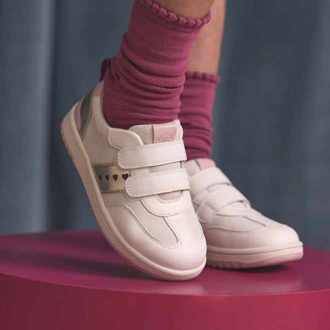 A child wears Pretty Brave River Hearts Leather Trainer Pink Off White with pink socks, standing on a round pink platform against a soft blue background. The shoes feature white genuine leather and small heart designs., 9420075363854, 9420075363847, 9420075363830, 9420075363823, 9420075363816, 9420075363809, 50502, 50503, 50504, 50505