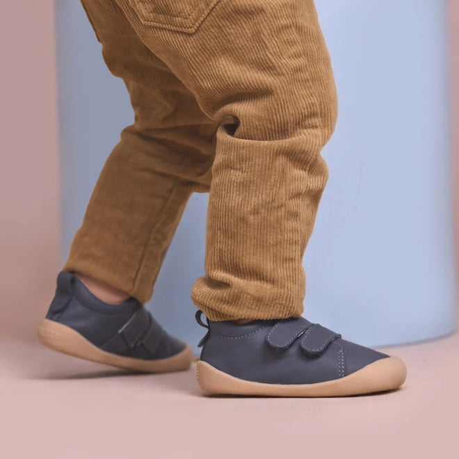 A toddler stands on a light pink floor against a pale blue and pink background, wearing tan corduroy pants and Pretty Brave First Walker Toto Leather Trainer Indigo Blue shoes with beige soles., 9420075366275, 9420075366268, 9420075366251, 50971