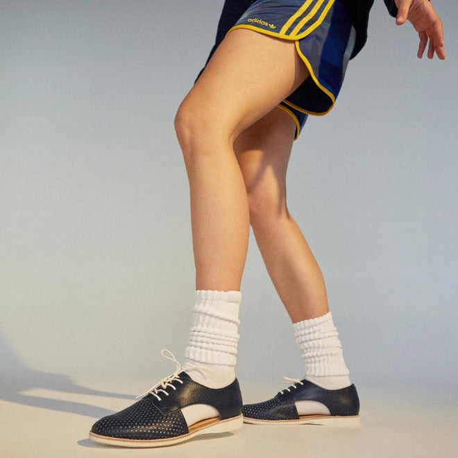 A person wearing navy and yellow athletic shorts, white socks, and Rollie Womens Sidecut Punch Navy Tumble Leather Shoes with anti-slip rubber outsoles and punch hole detailing steps forward against a neutral background., 9781760775414, 9781760775421, 9781760775438, 9781760775445, 9781760775452