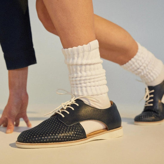 A person wears Rollie Womens Sidecut Punch Navy Tumble Leather Shoes with punch hole detailing and white laces, paired with thick white scrunched socks. One foot steps forward, ready to move—highlighting style and breathable linings., 9781760775414, 9781760775421, 9781760775438, 9781760775445, 9781760775452