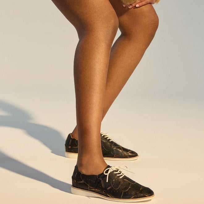 From mid-thigh down, a person with brown skin is shown bending slightly, wearing the Rollie Derby Applique Black Womens Leather Brogue Shoe—Australian designed, featuring black patterned leather uppers and white laces., 9781760792312, 9781760792329, 9781760792336, 9781760792343, 9781760792350