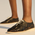 Close-up of a person wearing Rollie Derby Applique Black Womens Leather Brogue Shoes with cut-out soft leather uppers and beige soles, standing on a light surface. Only the lower legs and feet are visible., 9781760792312, 9781760792329, 9781760792336, 9781760792343, 9781760792350