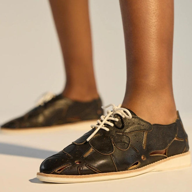 Close-up of a person wearing Rollie Derby Applique Black Womens Leather Brogue Shoes with cut-out soft leather uppers and beige soles, standing on a light surface. Only the lower legs and feet are visible., 9781760792312, 9781760792329, 9781760792336, 9781760792343, 9781760792350