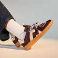 Wearing dark denim pants and white ribbed socks, a person rests one foot over the other, showcasing Rollie Womens Pace Dark Cherry Bone Suede Sneakers featuring a maroon and white suede upper, gum soles, and light blue laces., 9339554000315, 9339554000317, 9339554000164, 9339554000056, 9339554000182
