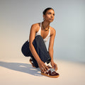 A woman crouches to tie her Rollie Womens Pace Dark Cherry Bone Suede Sneakers, styled with a white halter top, dark jeans, necklace, and rings, against a softly lit neutral background., 9339554000315, 9339554000317, 9339554000164, 9339554000056, 9339554000182