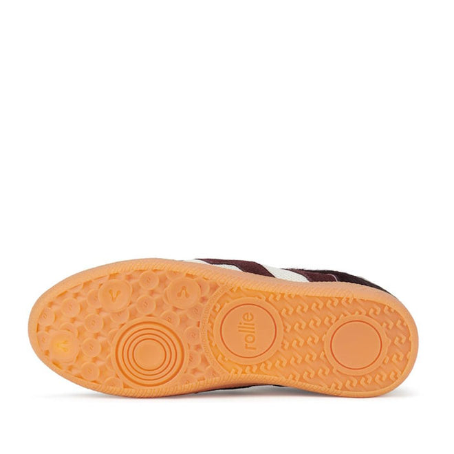 The image shows the orange rubber sole of the Rollie Womens Pace Dark Cherry Bone Suede Sneaker, featuring concentric circles, small raised dots for grip, and "rollie" embossed on its minimalist, textured design., 9339554000315, 9339554000317, 9339554000164, 9339554000056, 9339554000182