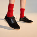 A person stands on a light surface wearing bright red socks and Rollie Womens Derby Black Splice Leather Brogue Shoes, featuring black lace-up design and anti-slip rubber outsoles. Only their lower legs and feet are visible., 51190, 9339554014920, 9339554054292, 9339554007458, 9339554000329, 9339554389769