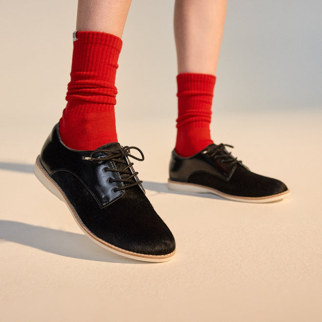 A person stands on a light surface wearing bright red socks and Rollie Womens Derby Black Splice Leather Brogue Shoes, featuring black lace-up design and anti-slip rubber outsoles. Only their lower legs and feet are visible., 51190, 9339554014920, 9339554054292, 9339554007458, 9339554000329, 9339554389769