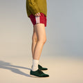 A person stands on a light-colored floor against a neutral background, wearing green shoes (Rollie Womens Derby Emerald Velvet Brouge Shoe), white socks, red shorts with white stripes, and a green sweater. Upper body and face are not visible. Outfit code: SC02101., 9339554068114, 9339554005690, 9339554000077, 9339554000336, 9339554023830