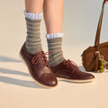 A person wearing gray striped socks with white ruffles and Rollie Womens Derby Super Soft Dark Cherry Leather Shoes stands on a light surface. A brown handbag with a green keychain is partially visible nearby., 9339554034843, 9339554086330, 9339554099231, 9339554070285, 9339554124001, 9339554126029
