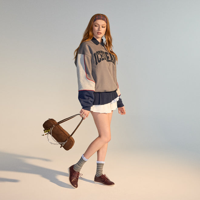 A young woman with long red hair wears a headband, an oversized ICECREAM sweatshirt, pleated white skirt, gray socks, and Rollie Womens Derby Super Soft Dark Cherry Leather Shoes. She holds a brown bag in a minimal, softly lit studio., 9339554034843, 9339554086330, 9339554099231, 9339554070285, 9339554124001, 9339554126029