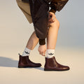 A person wearing Rollie Womens Chelsea Dark Cherry Leather Ankle Boots and white socks with black Adidas logos bends down to adjust one boot. Only the lower body is visible, styled with a brown skirt and shiny brown jacket (SC02136 look)., 9339554019543, 9339554104898, 9339554094687, 9339554000190, 9339554073989