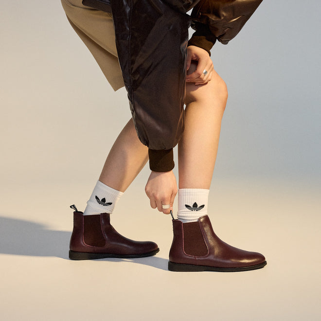 A person wearing Rollie Womens Chelsea Dark Cherry Leather Ankle Boots and white socks with black Adidas logos bends down to adjust one boot. Only the lower body is visible, styled with a brown skirt and shiny brown jacket (SC02136 look)., 9339554019543, 9339554104898, 9339554094687, 9339554000190, 9339554073989