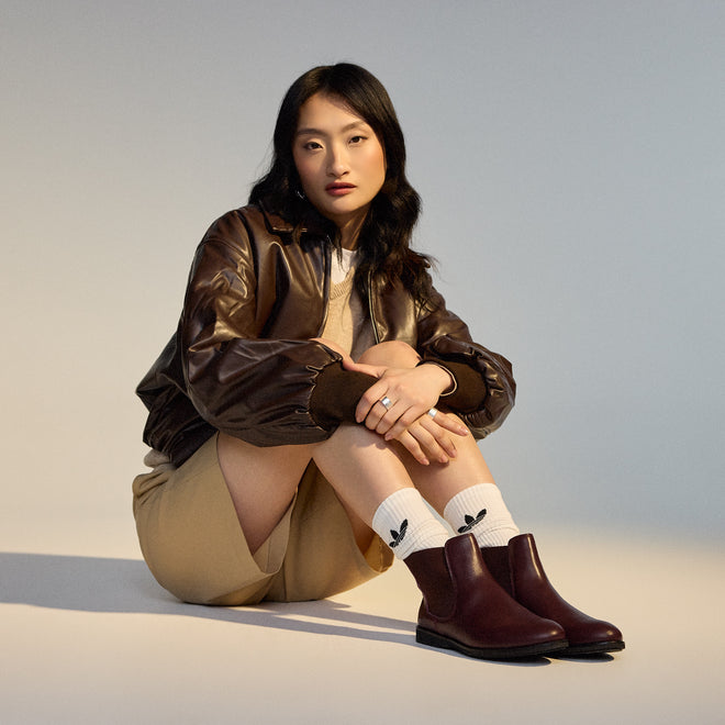 A woman with long dark hair sits on the floor, knees up, looking at the camera. She wears a brown leather jacket, beige shorts, SC02136 socks with black leaf logos, and Rollie Womens Chelsea Dark Cherry Leather Ankle Boot., 9339554019543, 9339554104898, 9339554094687, 9339554000190, 9339554073989
