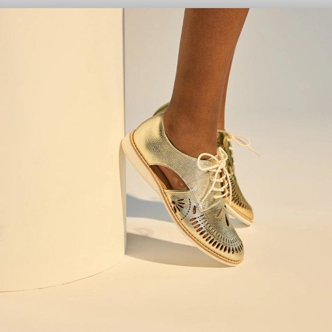 A person wearing Rollie Womens Sidecut Boho Punch Gold Leather Shoes, featuring cutout patterns and a memory foam footbed, stands on tiptoe beside a white cylindrical object., 9381760798811, 9381760798828, 9381760798835, 9381760798842, 9381760798859