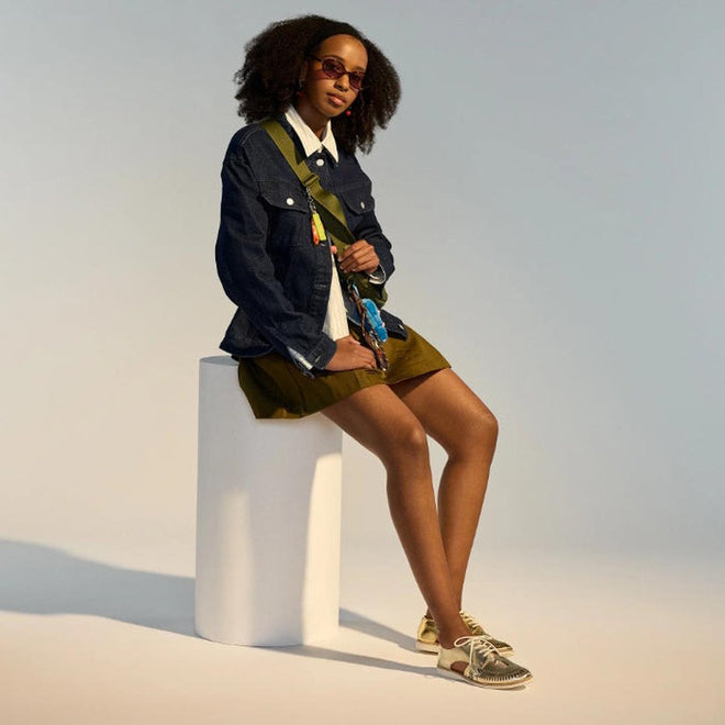 A woman with curly hair sits on a white stool wearing sunglasses, a denim jacket, white shirt, olive skirt, crossbody bag, and Rollie Womens Sidecut Boho Punch Gold Leather Shoes—all set against a neutral background., 9381760798811, 9381760798828, 9381760798835, 9381760798842, 9381760798859