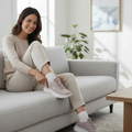 A woman sits on a light gray sofa, smiling as she puts on her Skechers Women's Slip-In Go Walk Arch Fit N-Joy Taupe shoes. Dressed in casual light-colored clothes, she relaxes in a modern, bright room with an abstract painting on the wall., 49941, 49942, 199025245340, 49944, 199025245364, 49946, 199025245388, 49948, 199025245401, 49950