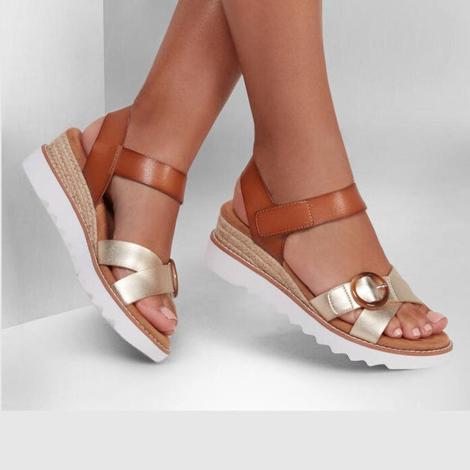A person wears Skechers Bobs Womens Desert Kiss Hi Divine Garden Champagne Tan sandals with tan and metallic gold straps, a decorative buckle, and white platform soles, standing against a light background., 195969581407, 40918, 195969581414, 40920, 195969581421, 40922, 195969581438, 195969581445, 40925