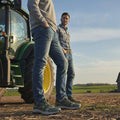 Two men in jeans and casual shirts stand outdoors by a green tractor on a farm. One wears Skechers John Deere Equalizer 5.0 Trail Harvester Slip Ins Green, with fields, a barn, and blue sky in the background., 50552, 197976615144, 197976615168, 197976615182, 197976615205