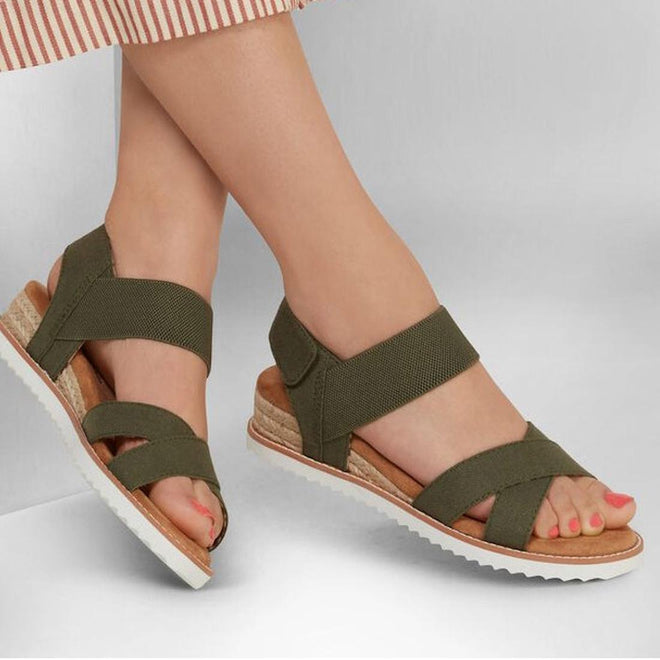 A person wears Skechers Bobs Womens Desert Kiss Secret Picnic Wedge Sandal in olive, featuring crisscross straps and a white ridged sole, paired with a striped skirt against a neutral background., 194428807249, 194428807256, 194428807263, 194428807270, 194428807287, 40840, 194428807300, 194428807317, 194428807225, 194428807201